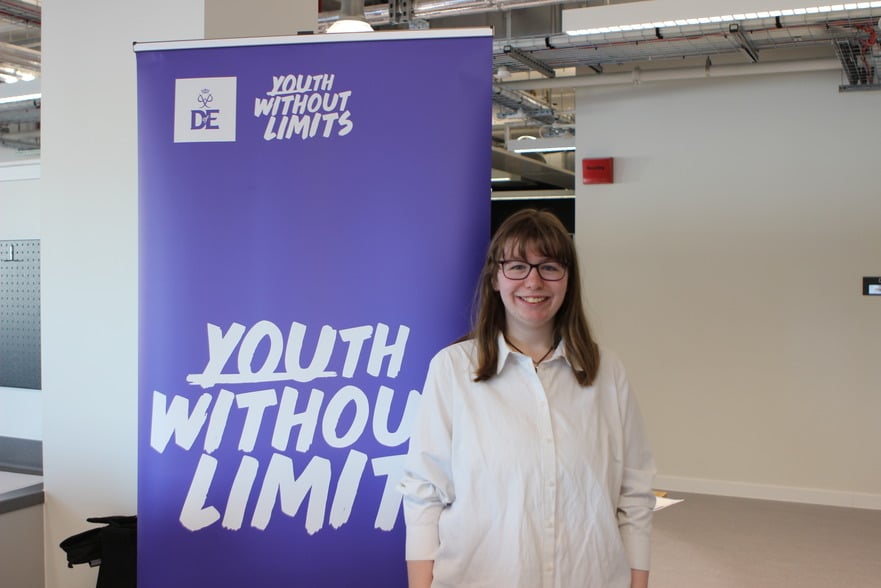 Jenny stands in front of a purple Youth Without Limits banner, smiling and wearing a white shirt.