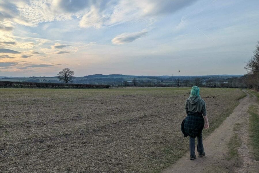 A person with teal-coloured hair walks along a dirt path beside an open, harvested field under a wide, cloud-streaked sky. They are wearing a hoodie with a checkered shirt tied around their waist. In the distance, rolling hills and scattered trees stretch toward the horizon as the sun begins to set.