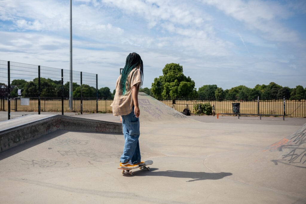 Amaris rides a skateboard at an outdoor skate park on a sunny day. They are wearing a beige plaid shirt, loose blue jeans, and orange sneakers. The skate park has ramps with graffiti, and a grassy field with trees is visible in the background beyond a metal fence.