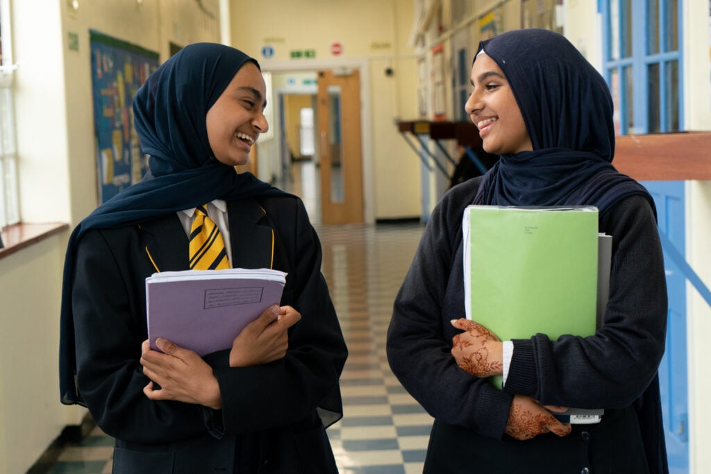 Zhaara and Maryam are standing in a school hallway, smiling at each other. One is holding a purple notebook and wearing a blazer with a yellow and black striped tie, while the other holds a green folder and has henna designs on her hands. They appear to be engaged in a friendly conversation.