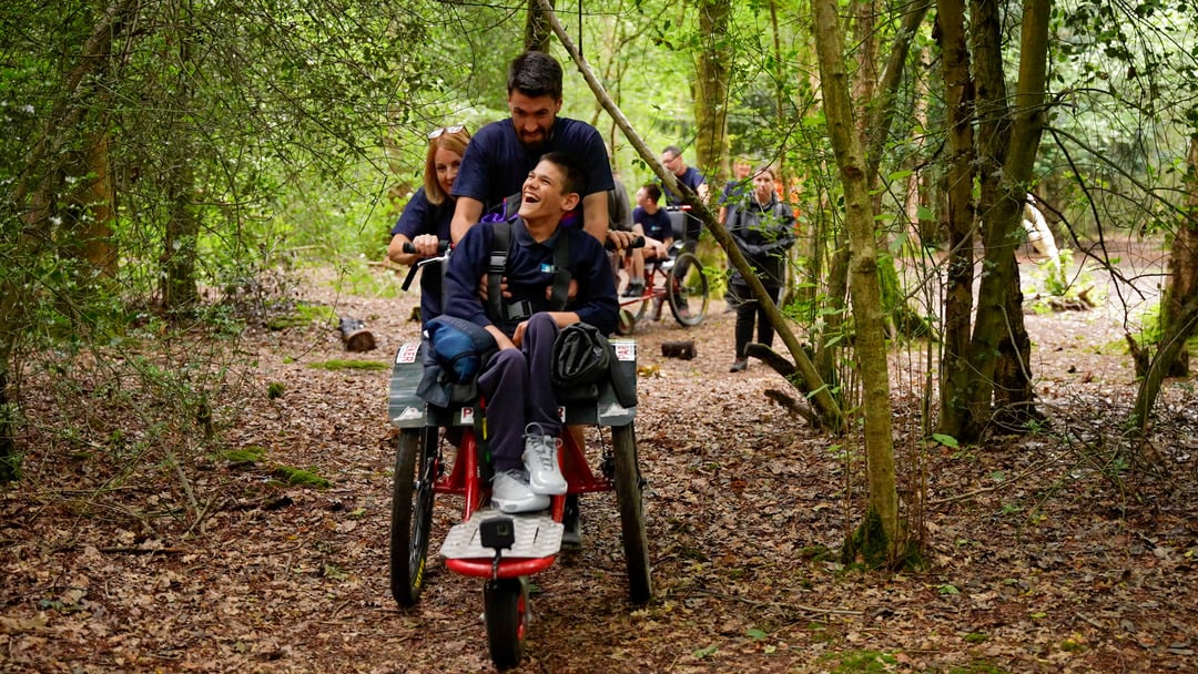 A group of people is walking in a forested area. One person is being pushed in a specialized wheelchair by another, both smiling and laughing. The surrounding trees create a lush, green backdrop, and other individuals can be seen in the background, also using similar wheelchairs.