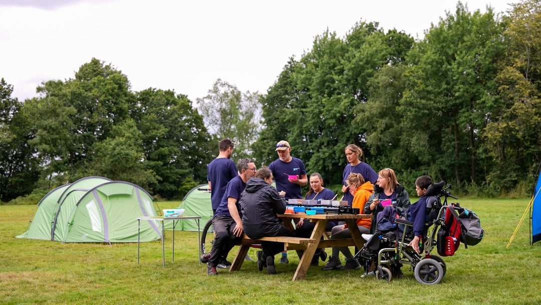 A group of people gathered around a picnic table in a grassy area, engaged in a board game. There are two green tents set up and trees lining the area.