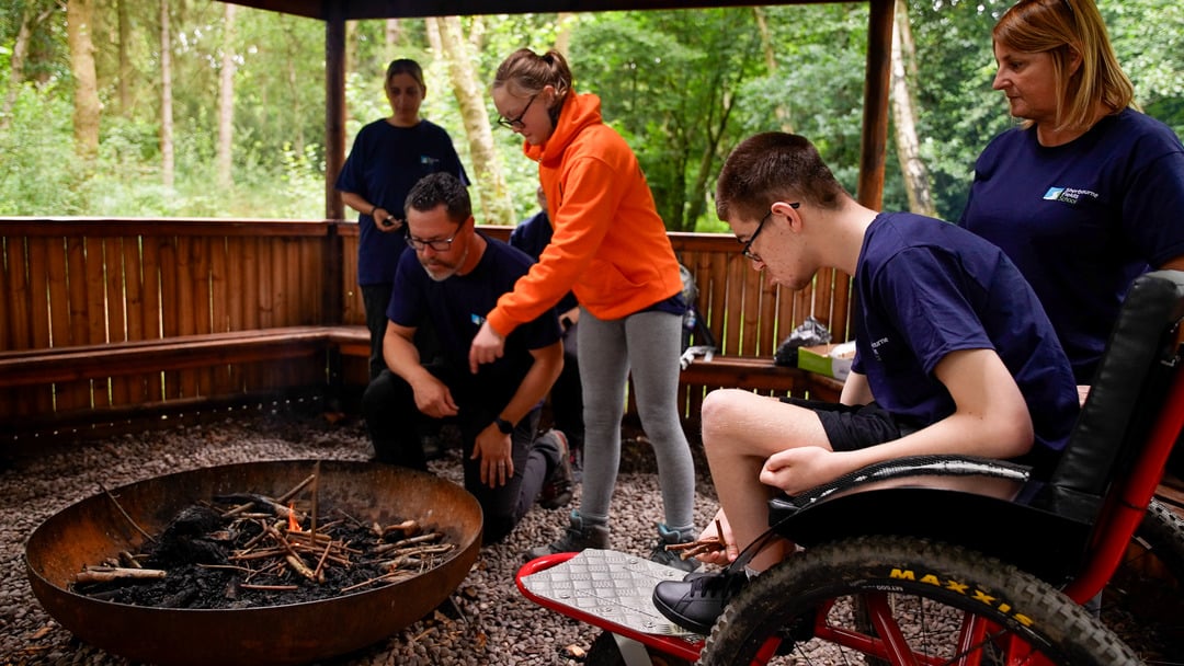A group of four people is gathered around a large fire pit in a sheltered, wooded area. One person in an orange hoodie is pointing at the fire pit, while a man in glasses kneels beside it. A young man in a wheelchair is sitting nearby, observing the scene. Another woman stands in the background, watching the group.