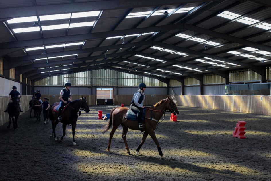 A group of riders on horseback are training indoors in a spacious arena. The arena has a high ceiling with skylights allowing natural light to filter in. The riders, dressed in riding gear and helmets, are guiding their horses along a sandy surface, with red cones placed in the arena for training purposes. The atmosphere is focused and calm, with wooden walls surrounding the space.