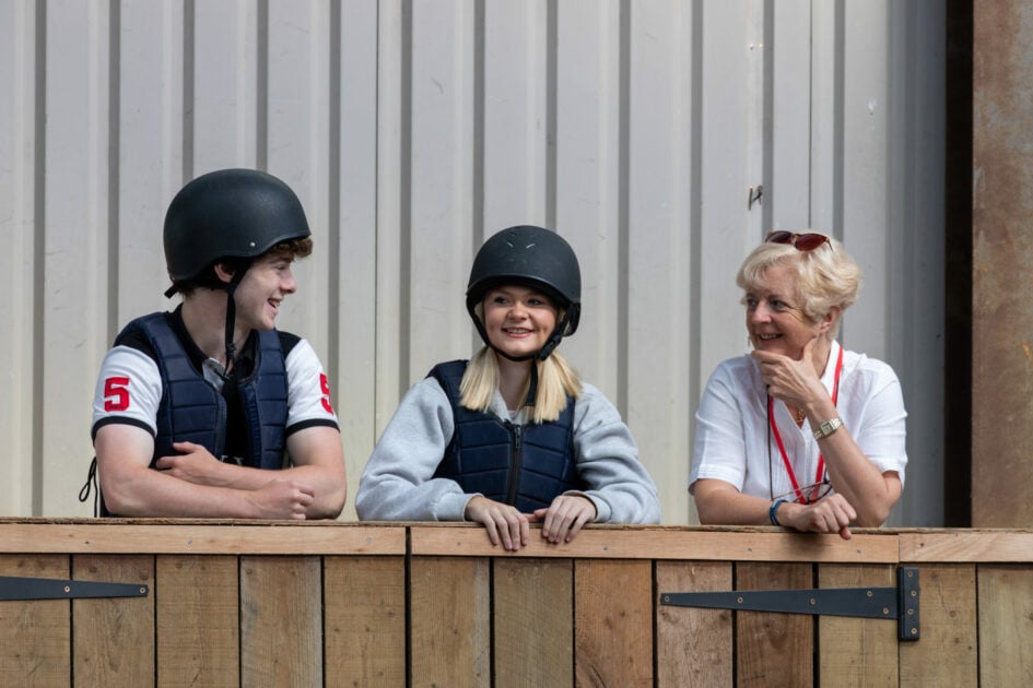 Three people standing by a wooden railing, wearing helmets and protective vests. Two young people are smiling and engaging in conversation, while an older woman, also smiling, listens attentively.