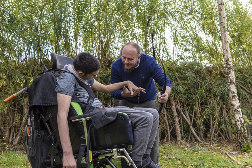 A young man in a wheelchair sitting outdoors, looking down at his hands. A man beside him leans in, gently holding his hand and engages with him.