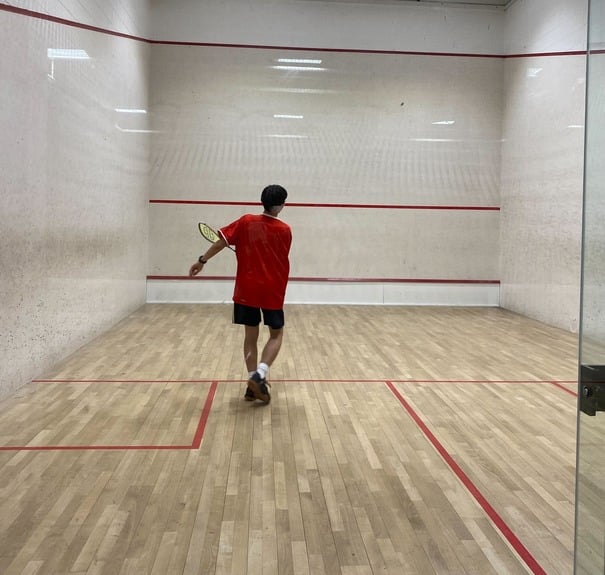 Evan in a red t-shirt and black shorts playing squash inside a court.
