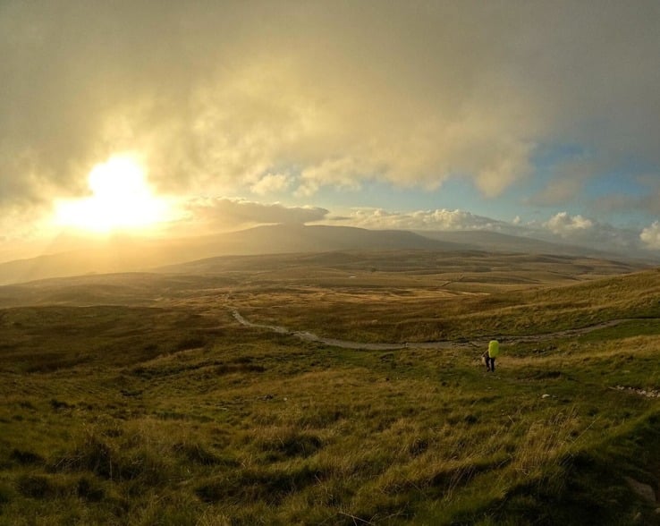The sun rising over grassy hills, with one person walking on a path.