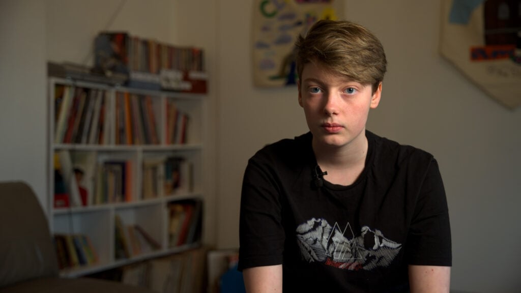 Ellesha has short light-brown hair and a black T-shirt, sits indoors in front of blurred bookshelves, looking calmly at the camera.