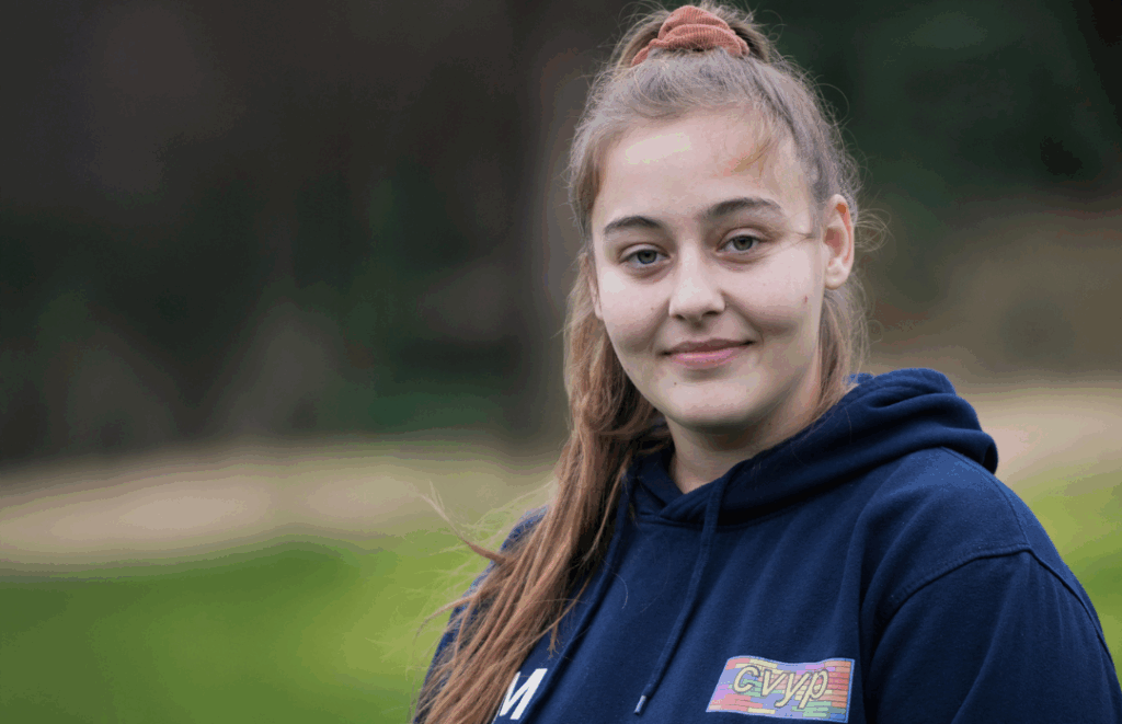 Sophie is standing outdoors, smiling softly at the camera. She is wearing a navy hoodie with a colourful "CVYP" logo on the chest. The background is grassy with blurred trees.