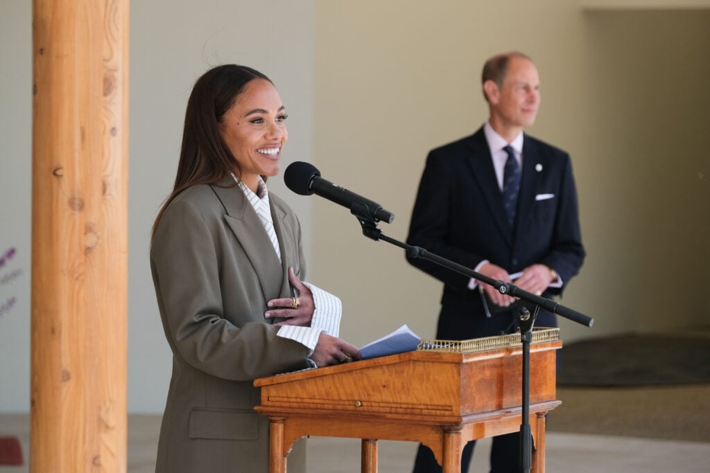 Alex Scott MBE smiles as she speaks at a podium with a microphone on the West Terrace at Buckingham Palace. She is wearing a khaki blazer over a striped shirt. In the background, HRH The Duke of Edinburgh stands slightly out of focus, wearing a dark suit and tie. The setting is formal and elegant, with wooden architectural features and a polished wooden podium.