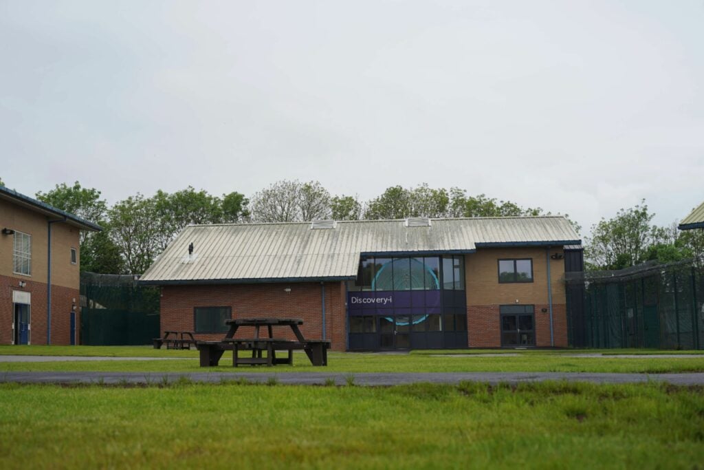 A modern, low-rise brick and metal building with a sign reading "Discovery-i" on the front. The building has a sloped metal roof and large windows. In the foreground, there are wooden picnic benches on a grassy lawn. The area is enclosed by fencing, with additional buildings visible to the left and right. The sky is overcast.