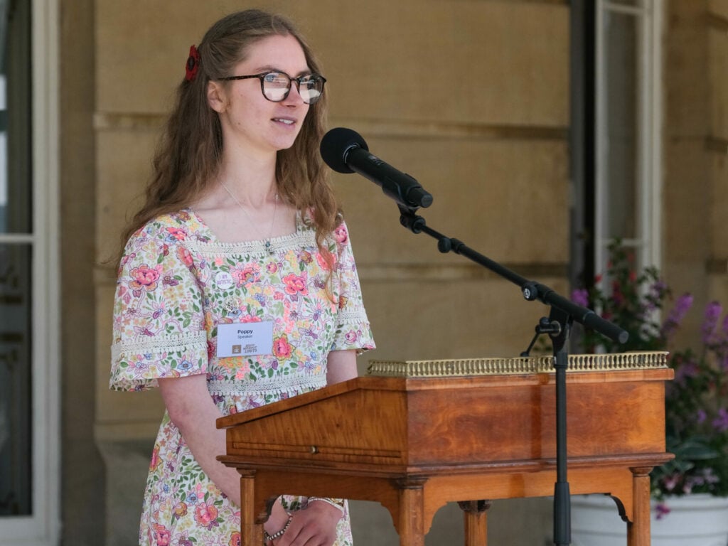 Poppy stands at a wooden podium with a microphone, speaking confidently. She is wearing glasses and a light-colored dress with a floral pattern. The background shows part of a grand building with stone walls and potted flowers, suggesting a formal outdoor setting.