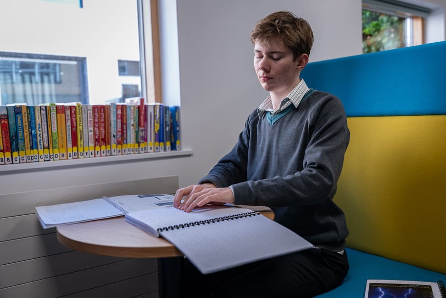 A person in school uniform sitting in the library reading Braille.