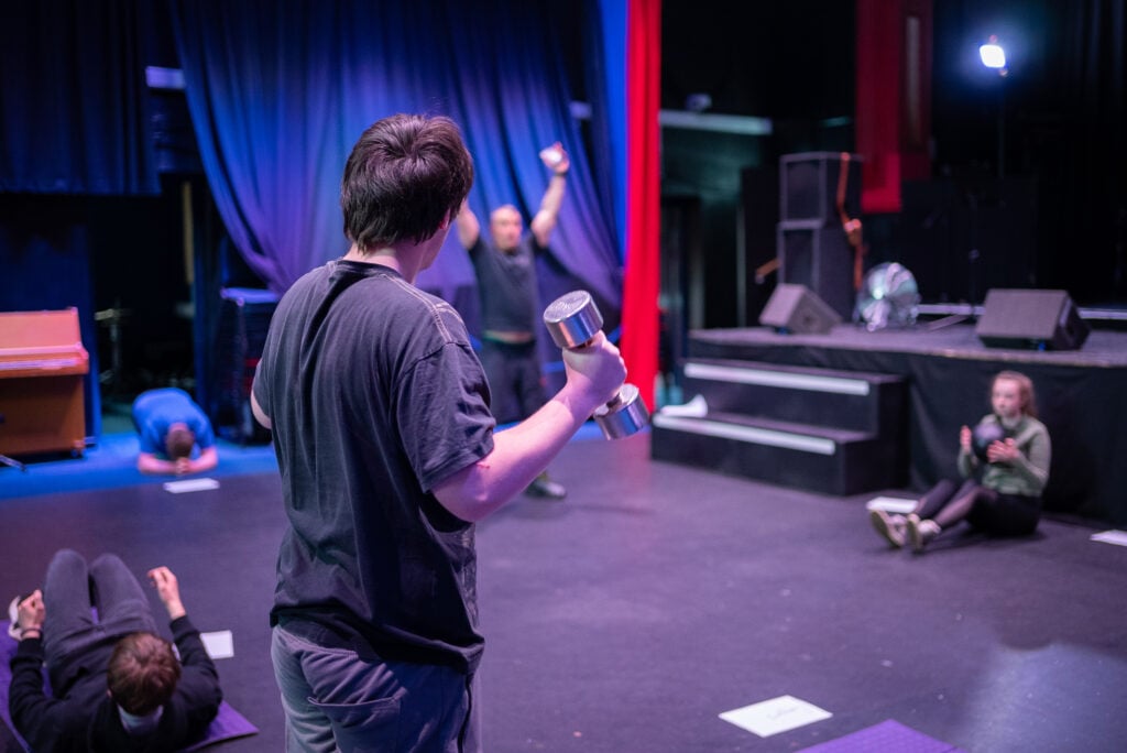 Lewis holding dumbbells, looking at Barry doing an exercise.
