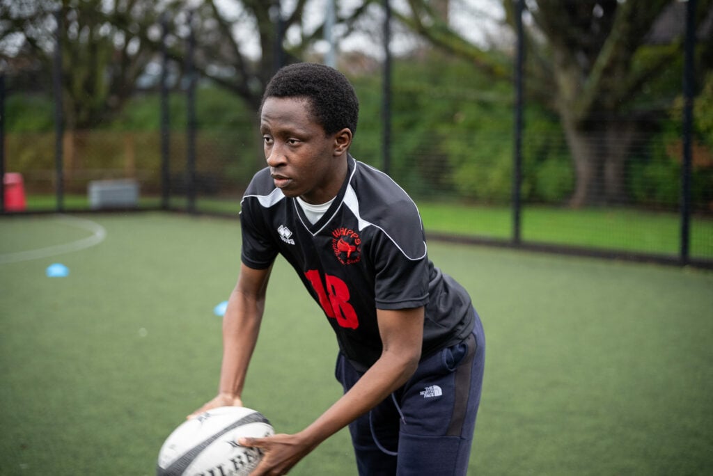 A young male athlete in a black sports jersey with the number 18 is preparing to pass a rugby ball on a grassy field. He appears focused and determined, with trees in the background and training cones scattered around the field.