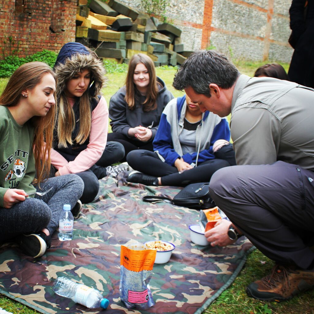 David Love kneels on a camouflage picnic blanket during a DofE expedition, preparing food from a packet while a group of teenage girls sit around him, watching attentively and smiling. The group is gathered in a grassy outdoor area beside old brick and stone walls, with foam blocks stacked nearby—creating an informal and supportive setting for the expedition activity.