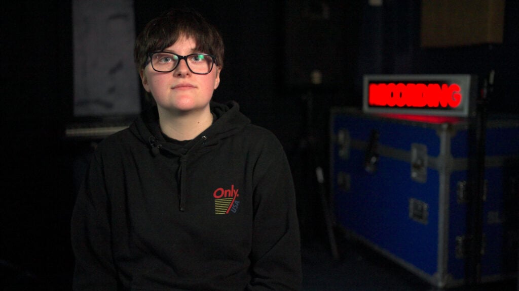 Jay, a person with short dark hair and glasses, is sitting in a dimly lit recording studio wearing a black hoodie that says “Only USA.” Behind him, a red “RECORDING” sign is illuminated on top of a large blue equipment case.