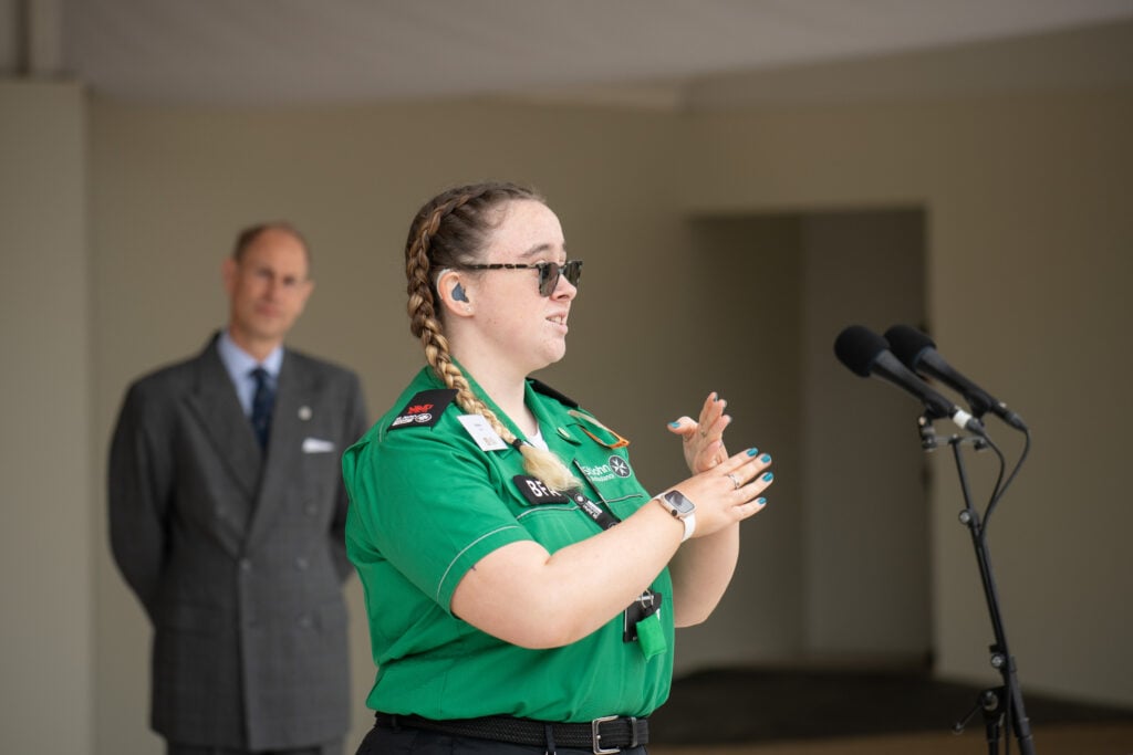 Hafwen, a young woman in a green St John Ambulance uniform, stands at a microphone signing in British Sign Language. She wears glasses, has braided hair, and an Apple Watch on her wrist. A man in a suit stands in the background watching attentively.