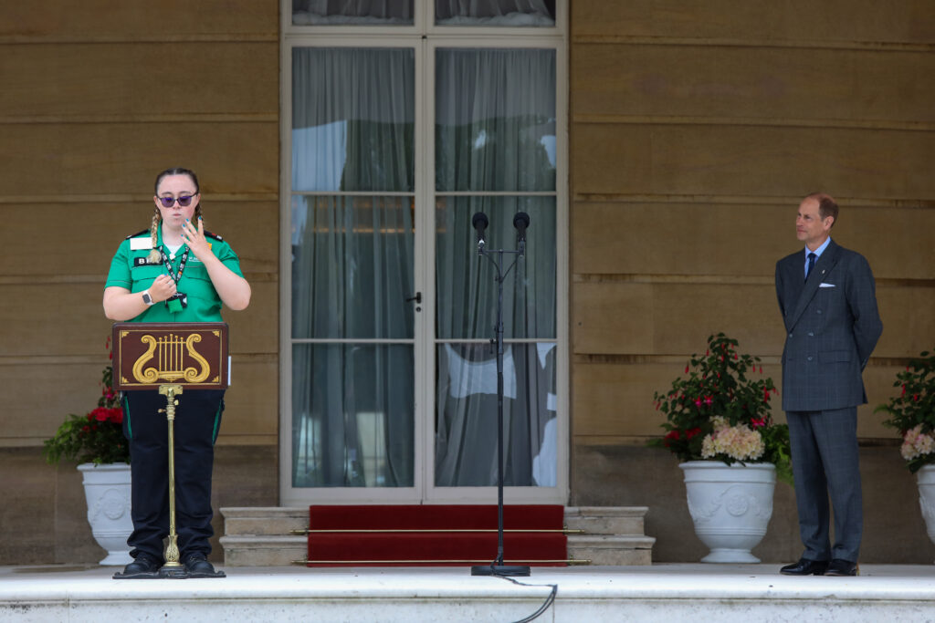 Hafwen, wearing a green St John Ambulance uniform and sunglasses, stands at a podium on the steps of a grand building, signing in British Sign Language. To her right, HRH The Duke of Edinburgh, dressed in a grey double-breasted suit, stands watching respectfully. They are outside a large set of glass doors, flanked by white flowerpots with red and white flowers.