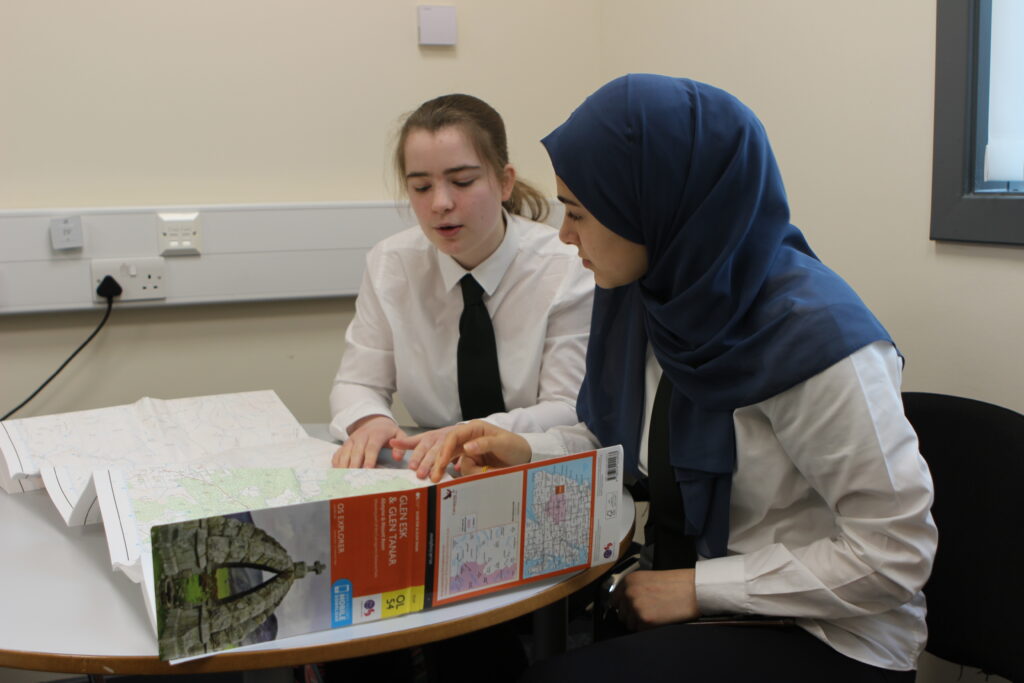 Two individuals sit at a table examining a large map. One wears a white shirt and black tie, the other a white shirt and blue headscarf, with an open paper map sitting on the table in front.