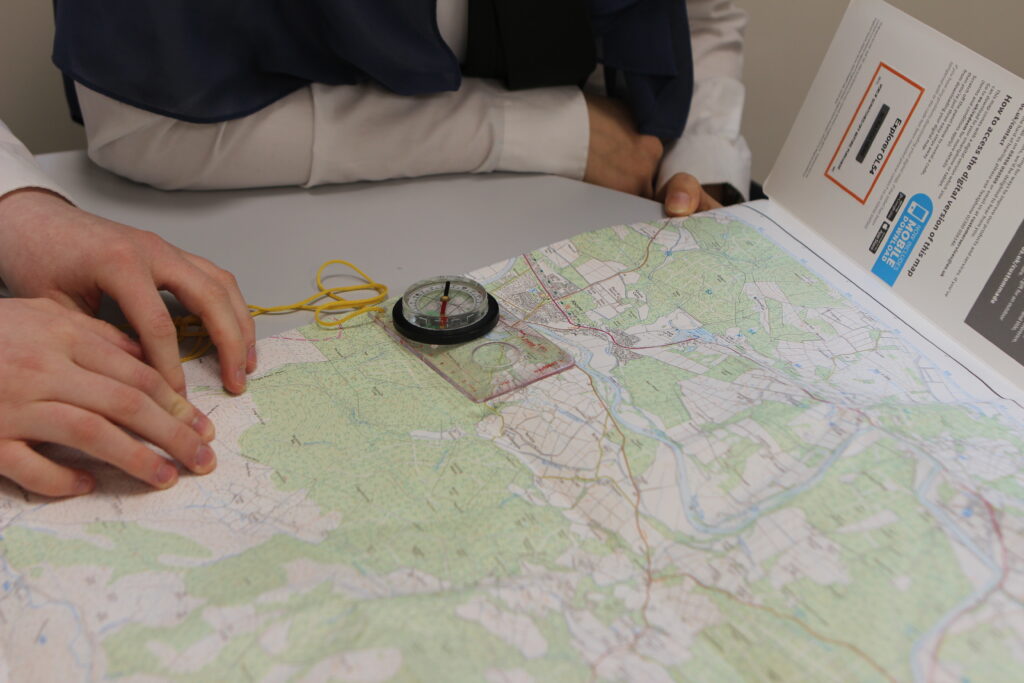 Two people sit at a table with a map spread out in front of them. One holds the map while the other places a hand on it. A compass rests on the map, and an open booklet with text and images lies to the right