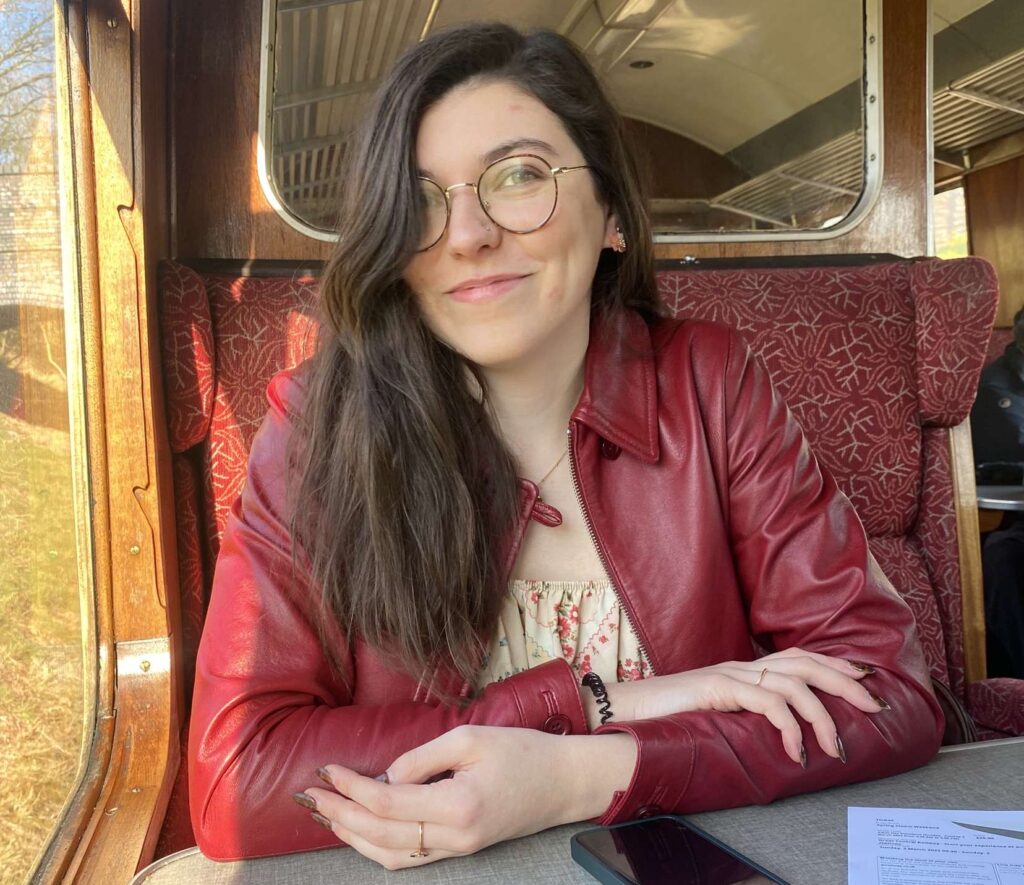 Charlotte sits at a table inside a train car. She is wearing a red leather jacket over a light-colored top and is smiling at the camera. Sunlight is streaming in through the window, illuminating her face and the cozy interior of the train. A smartphone and a piece of paper are on the table in front of her.