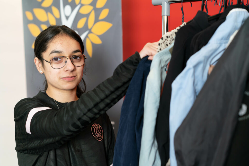 Zainab wearing glasses and a dark long-sleeve shirt stands beside a clothing rack, organizing garments. A banner with a yellow-leaf tree design is visible in the background.