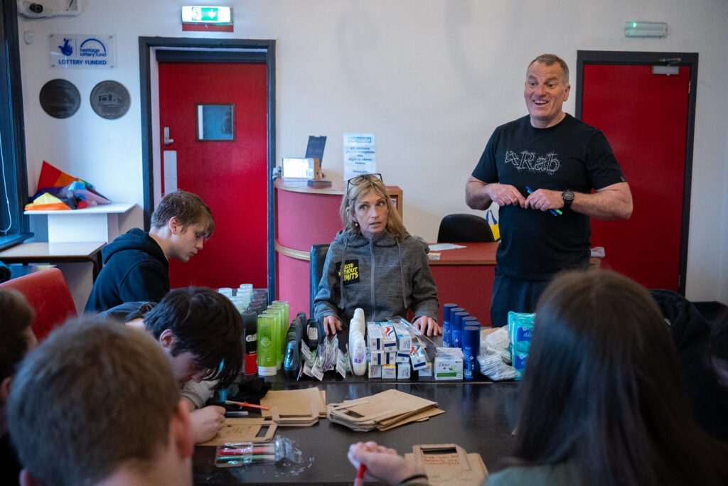 A group of people in a community center assembling care packages with hygiene products like toothpaste, toothbrushes, soap, and paper bags. Barry stands giving instructions while others are seated and engaged in the activity.