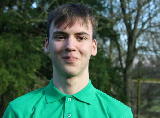Ellis has dark blonde hair and a green shirt, standing in front of green trees.