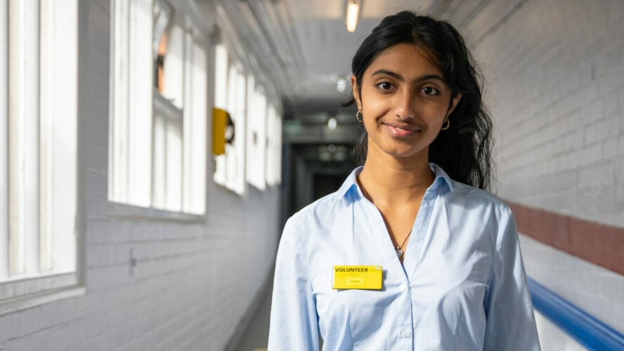 Dora has black hair and a blue shirt, standing in a hallway.