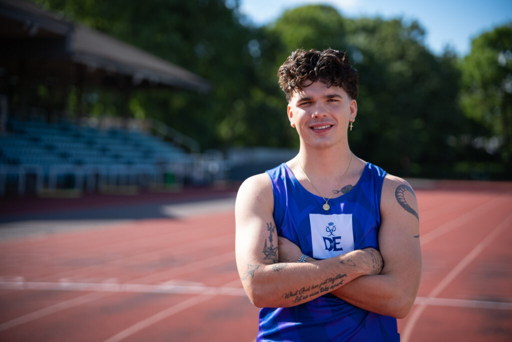 Harry Clark standing confidently on a running track with arms crossed, wearing a blue tank top featuring the letters 'DofE' and an emblem. The background includes empty stadium seating and trees. The athlete has tattoos on both arms and is smiling at the camera.
