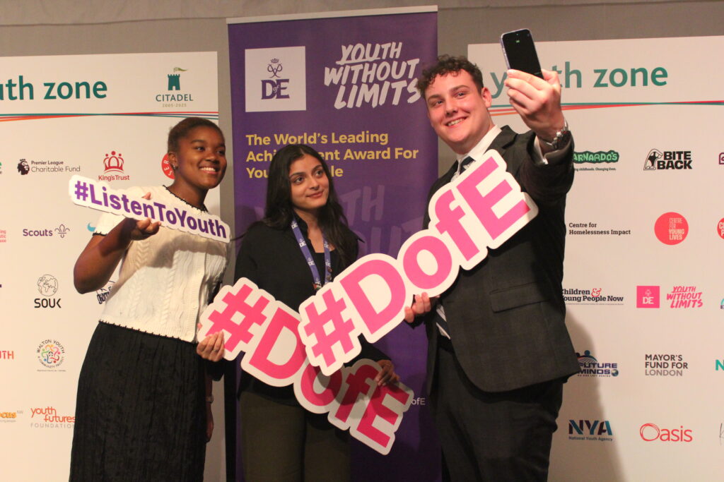 Three young people holding signs that say D of E and Listen to youth, one of them is taking a selfie.