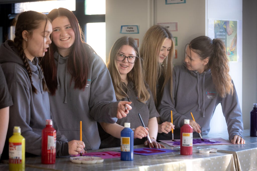 A group of five young women wearing gray hoodies are engaged in a painting activity. They are standing around a table covered with art supplies, including bottles of paint in red, yellow, and blue. The women are smiling and interacting with each other as they work on their projects, which appear to involve colorful designs. The background features a bright, well-lit room with art-related materials displayed on the walls.