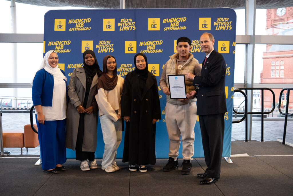 HRH The Duke of Edinburgh stands on a stage with five young people in front of a backdrop displaying the Duke of Edinburgh’s Award branding and the slogan “Youth Without Limits” in English and Welsh. One person is holding a framed certificate while the Duke gestures toward it. Large windows behind the group reveal part of a historic red-brick building outside.