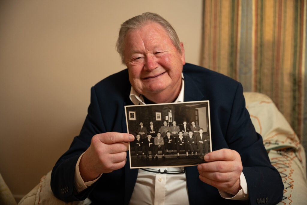 John Green sitting on a white patterned armchair against a neutral background, wearing a dark suit jacket over a light shirt, and holding up a black and white photograph of a group of boys smiling to camera.