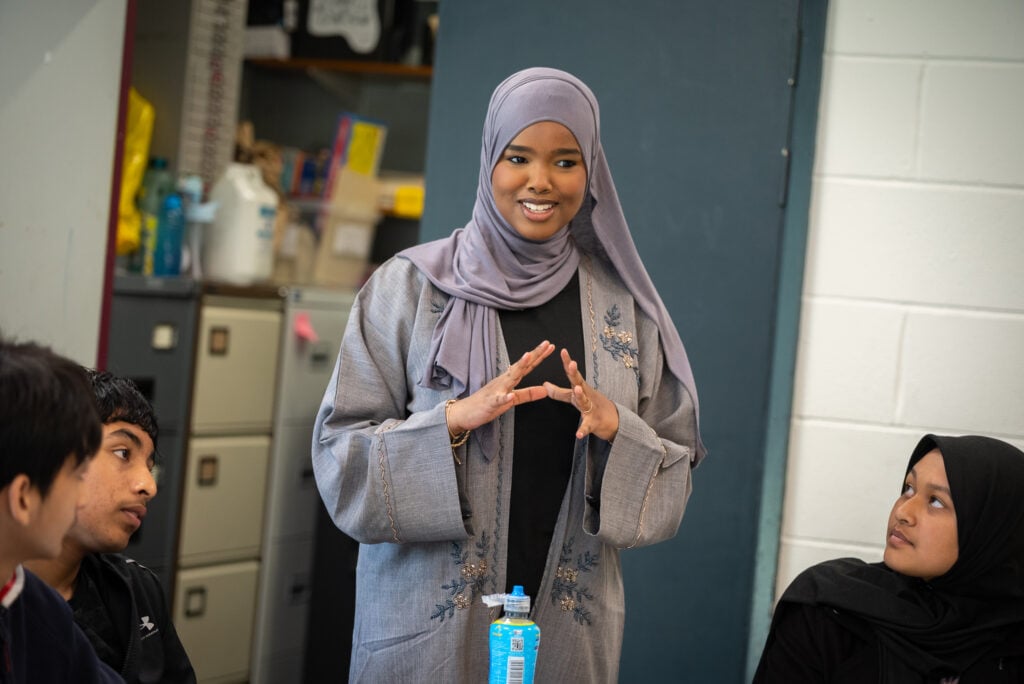 Amira standing up and talking to three people sitting down, she is wearing a purple dress and hijab.