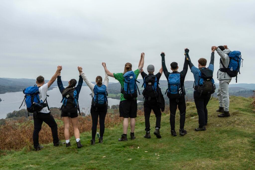 Eight hikers wearing backpacks stand on a grassy hilltop holding hands and raising their arms in celebration facing a scenic view.