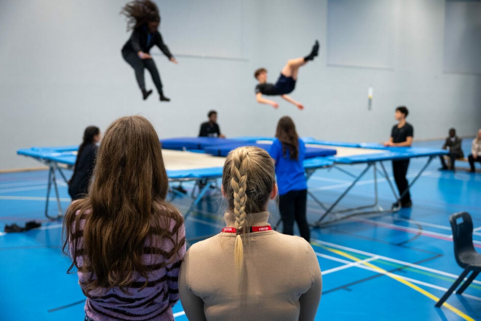 Two people looking at two DofE participants showing off their trampolining skills inside a sports hall.