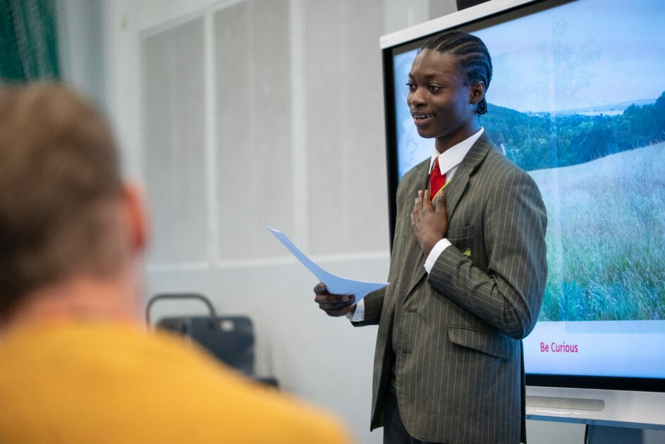 A pupil wearing a gray striped suit jacket and red tie presenting in front of a screen.