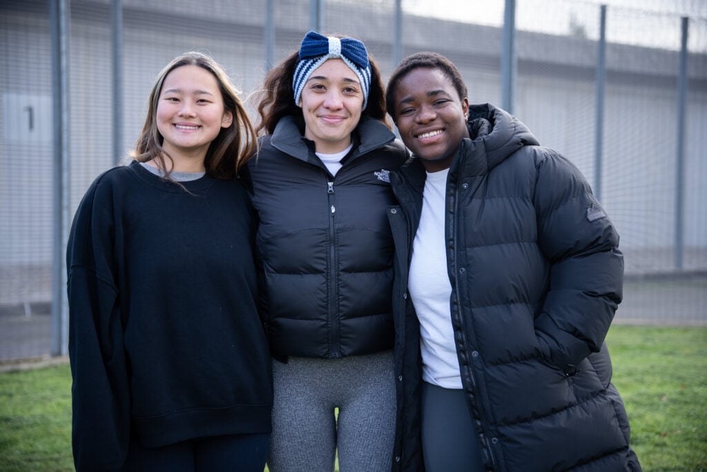 Three young women stand together outdoors, smiling at the camera. They are dressed in casual attire, with one wearing a black sweatshirt, another in a puffy black jacket, and the third in a gray top and leggings. The background features a fenced area, suggesting a sports or recreational setting.