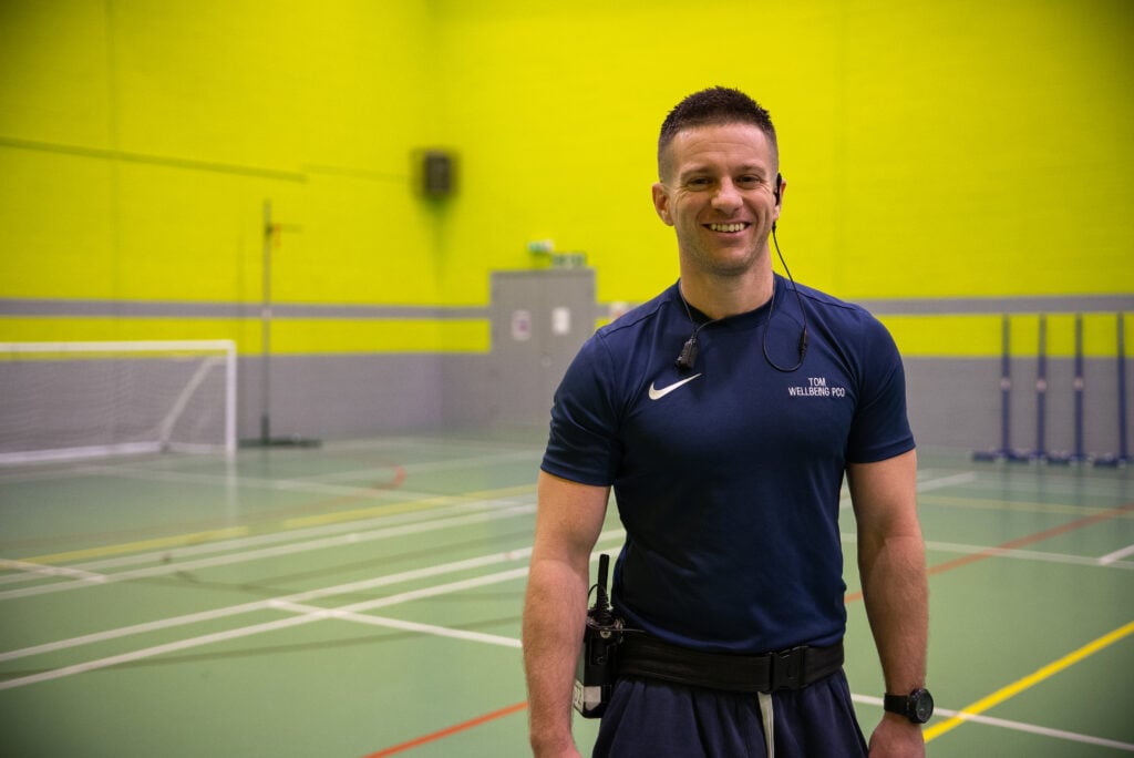 A smiling male fitness instructor stands in a brightly lit indoor sports facility. He is wearing a navy blue athletic shirt and has a headset and a radio attached to his waist. The background features a green wall, a soccer goal, and various sports equipment.