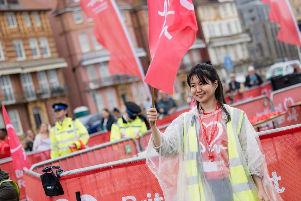 A person wearing a high‑visibility vest and a clear rain poncho stands beside event barriers, waving a large red British Heart Foundation flag. Several other flags and volunteers, along with police officers in yellow jackets, are visible in the background on a street lined with older buildings.