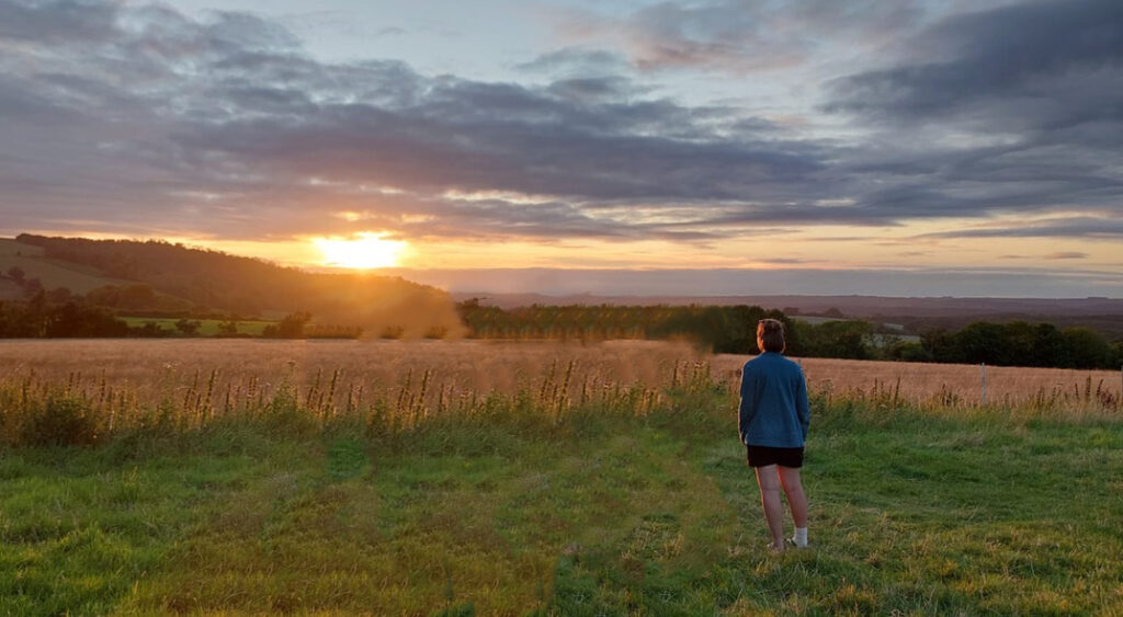 Anais standing in a grassy field looking at the sun set behind fluffy, blue clouds.