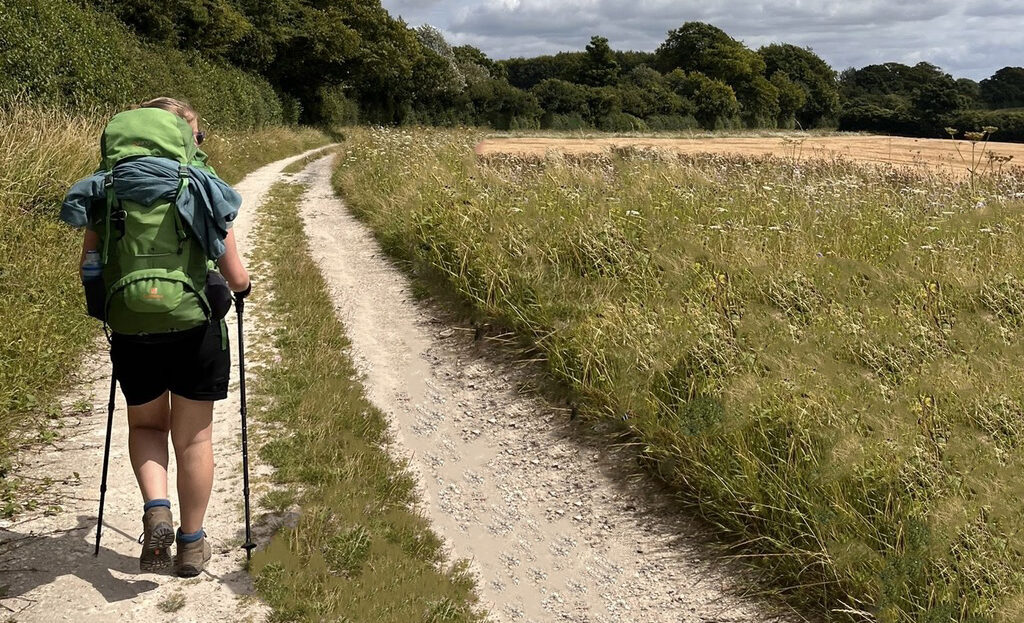 Anais carrying a green backpack and using walking poles down a gravel path.