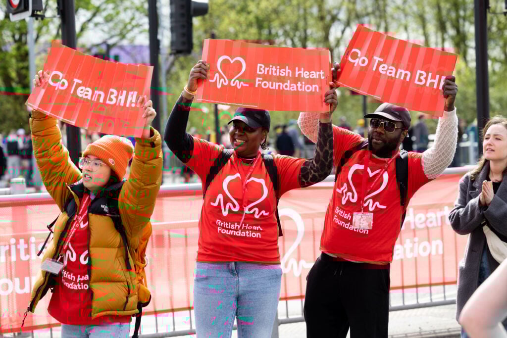 Three people wearing bright red British Heart Foundation T-shirts stand cheering behind a roadside barrier at an outdoor event. They hold up red signs that read “Go Team BHF!” and “British Heart Foundation.” Other spectators stand nearby, and trees and metal barriers line the background.