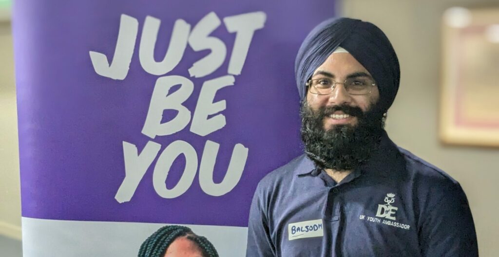 Baljodh wearing a D of E Youth Ambassador polo shirt standing in front of a banner that says "just be you".