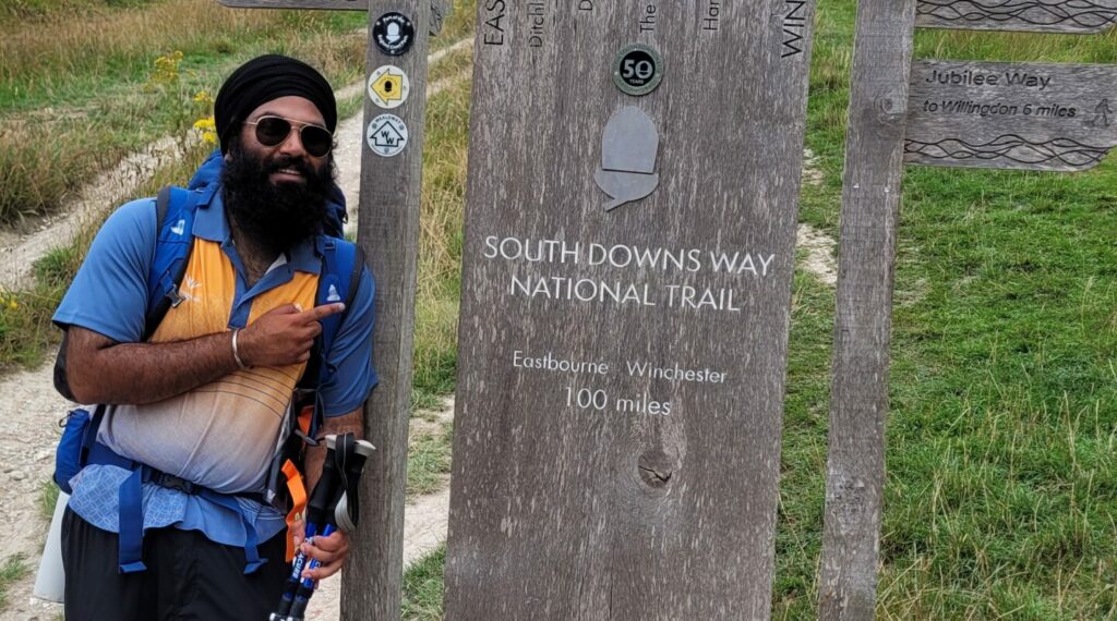 Baljodh wearing a blue and orange tshirt and holding walking poles pointing at a sign that says "South Downs Way National Trail".