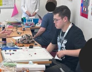 A group of young people are gathered around tables in a classroom, working on creative craft and film‑related projects. The tables are covered with materials including cardboard, tape, paper, markers, and small props. Some participants are standing while others are seated, busy assembling or designing items. The classroom walls are decorated with posters, artwork, and notices.