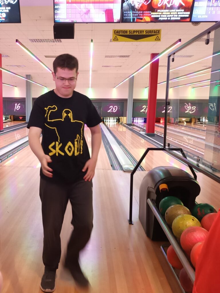 A person wearing a black T‑shirt with a yellow graphic of a flexing figure and the word “SKOL” stands on a bowling lane walkway. Bowling balls in various colors sit in a rack to the right, and multiple lit bowling lanes stretch out behind them. Overhead screens and a yellow “Caution Slippery Surface” sign are visible above the lanes.